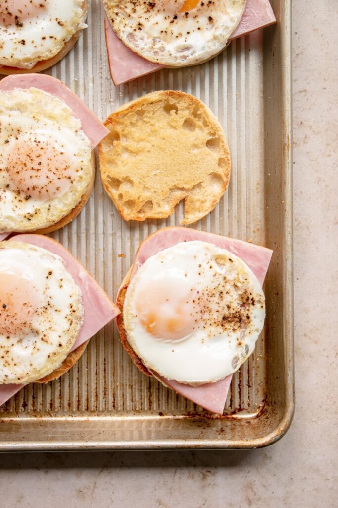 English muffins on a sheet pan with ham and cooked eggs