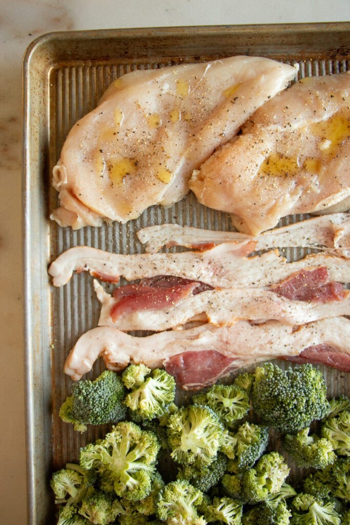 raw chicken breasts, bacon, and broccoli on a sheet pan with olive oil and pepper
