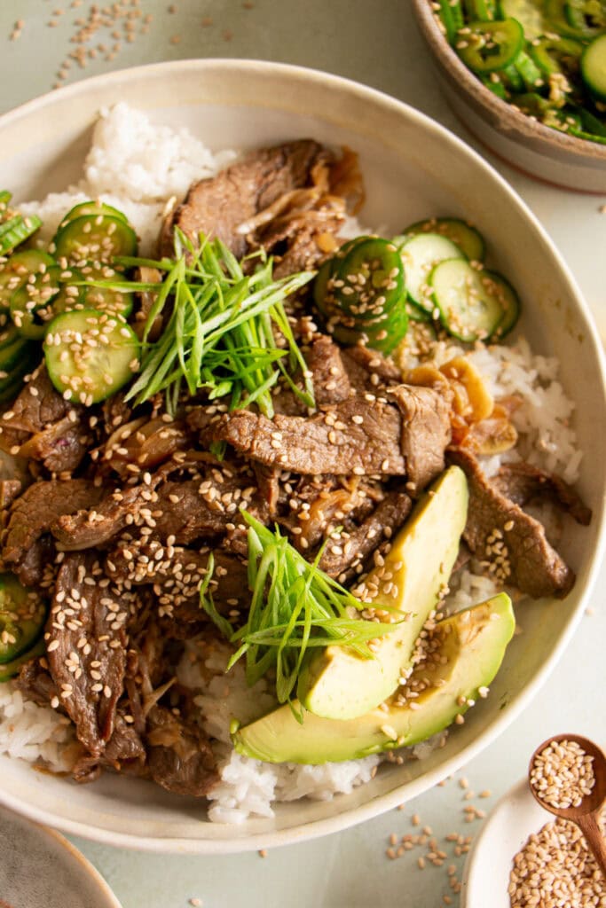 spicy sheet pan beef over rice with green onions, sesame seeds, cucumber salad, and avocado.