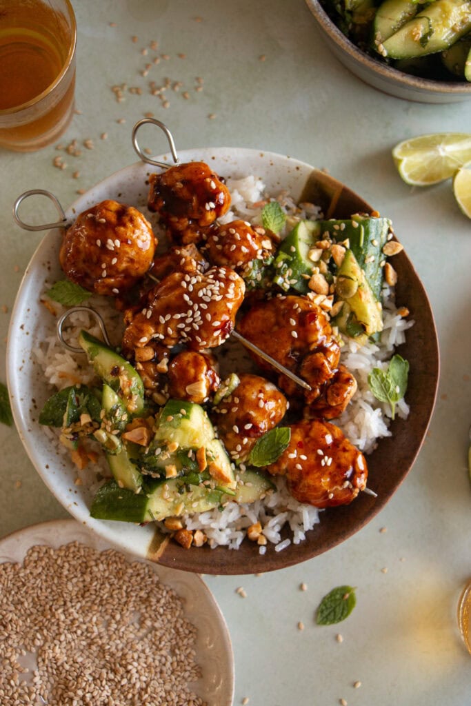 bowl of rice with general tsos chicken skewers and cucumber salad 