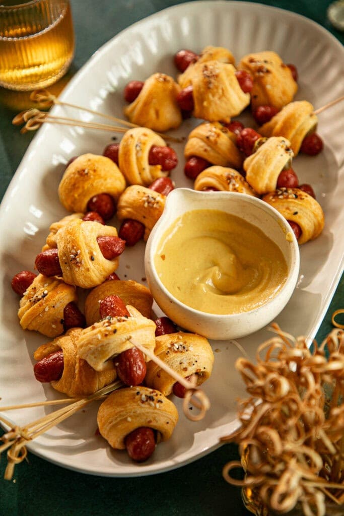 Large platter with pigs in a blanket and wooden appetizer picks and a bowl of mustard.