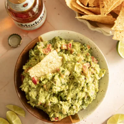 Overhead shot of a bowl of chunky guacamole with a tortilla chip dipped in, surrounded by lime wedges, chips, and Mexican beer
