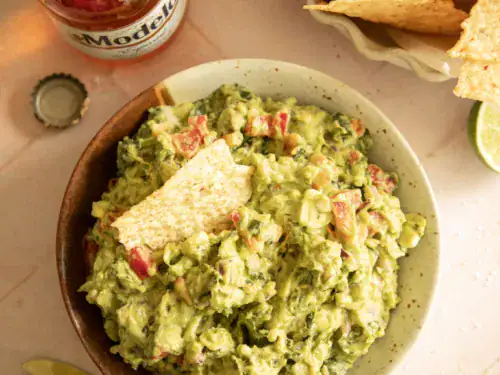 Overhead shot of a bowl of chunky guacamole with a tortilla chip dipped in, surrounded by lime wedges, chips, and Mexican beer