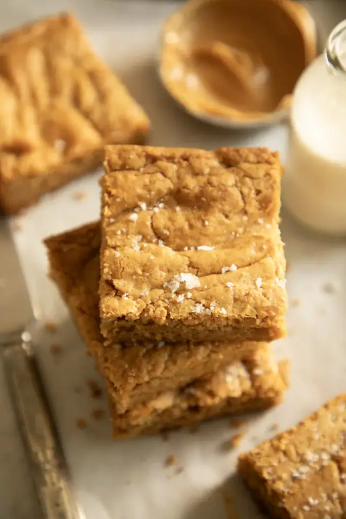 Close-up of fudgy peanut butter cookie bars stacked with a bottle of milk and a dish of peanut butter in the background