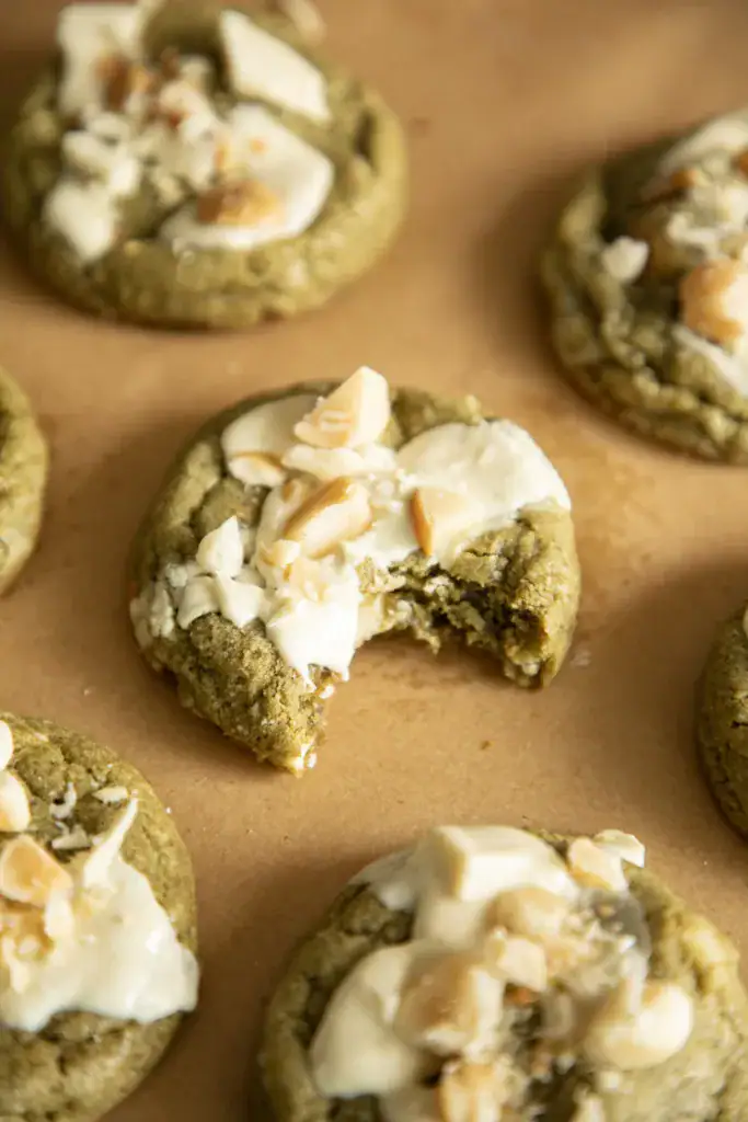 Matcha cookies on parchment paper, topped with white chocolate, one with a bite taken out.