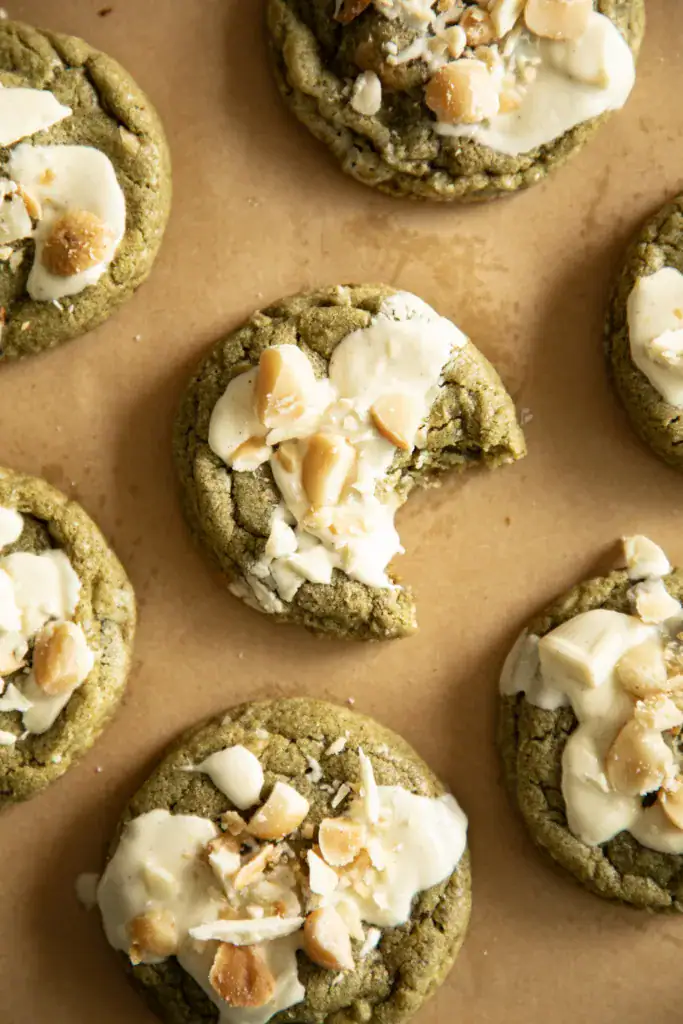 Overhead view of matcha cookies with white chocolate and macadamia nuts on parchment, one with a bite taken out