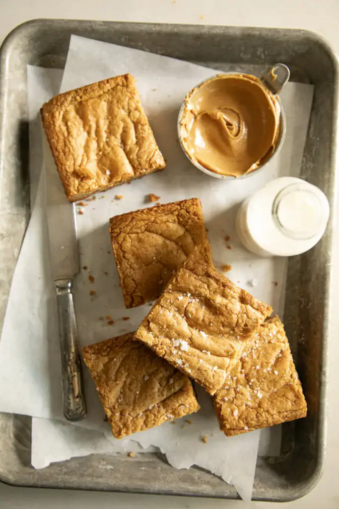 Overhead shot of peanut butter cookie bars on a metal baking tray with a small jar of milk and a bowl of peanut butter