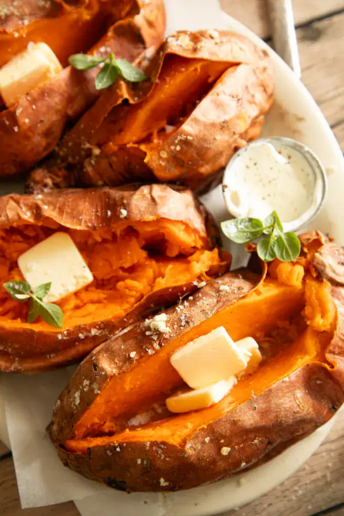 Close-up of grilled sweet potatoes topped with butter, with a cup of sour cream and fresh herbs.