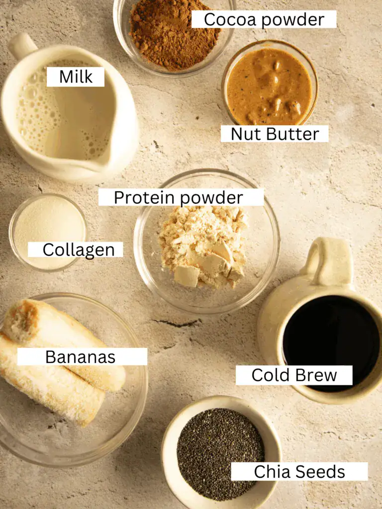 Top-down view of labeled smoothie ingredients including milk, cocoa powder, nut butter, bananas, cold brew, chia seeds, protein powder, and collagen.