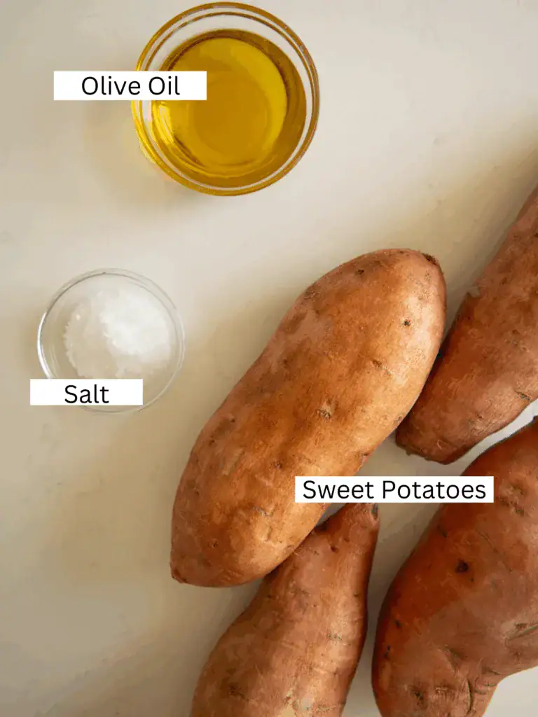 Overhead photo of raw sweet potatoes, olive oil, and flaky salt arranged on a light surface.