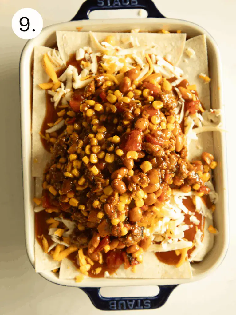 Tortilla layers topped with chorizo, corn, beans, tomatoes, and enchilada sauce inside a baking dish.