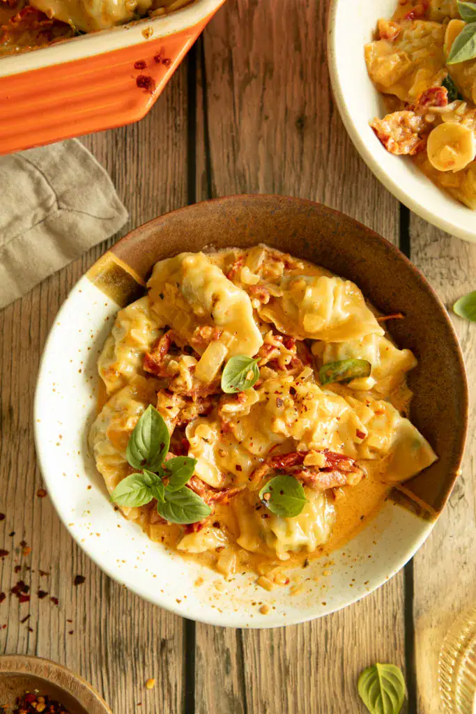 Two bowls of creamy baked dumplings on a wooden table, styled with basil and chili flakes.