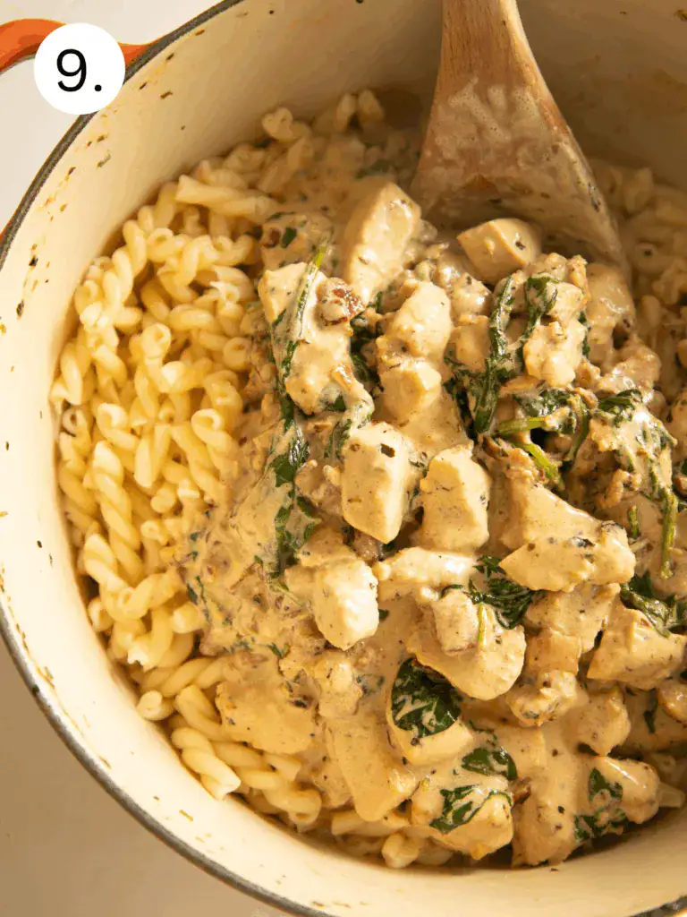 Creamy chicken, spinach, and bacon sauce being combined with cooked pasta in a Dutch oven.