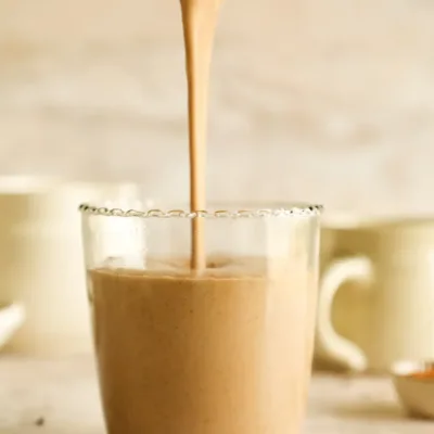 Chocolate smoothie being poured from a blender into a glass with blurred mugs in the background.