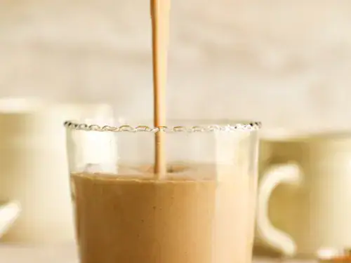 Chocolate smoothie being poured from a blender into a glass with blurred mugs in the background.
