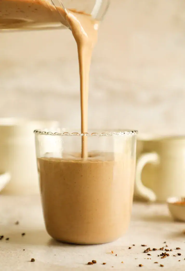 Chocolate smoothie being poured from a blender into a glass with blurred mugs in the background.