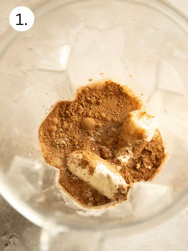 Cocoa powder, frozen banana, and protein powder in a blender before blending.