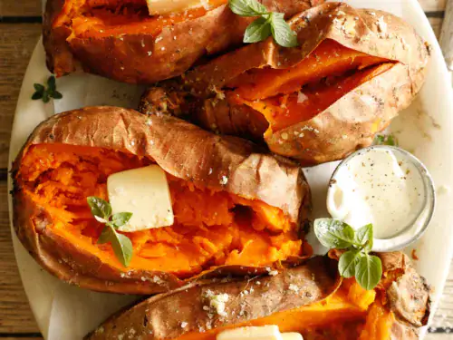 Overhead of grilled sweet potatoes opened and topped with butter, surrounded by herbs and drinks.