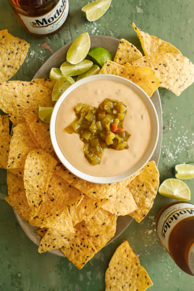 Bowl of creamy queso dip topped with green chiles, served with tortilla chips and lime wedges.