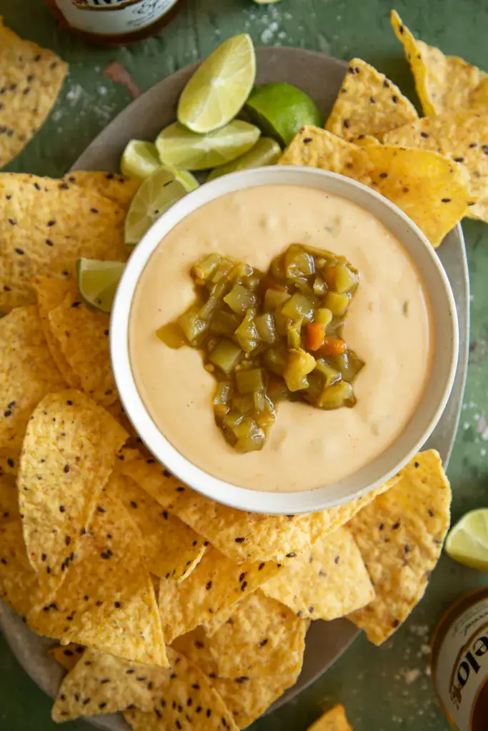 Overhead shot of queso dip surrounded by chips, lime wedges, and bottles of Modelo beer.