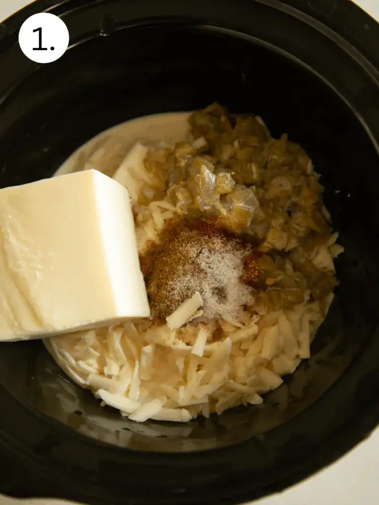 Shredded cheese, Velveeta, green chiles, spices, and milk in a slow cooker before cooking.