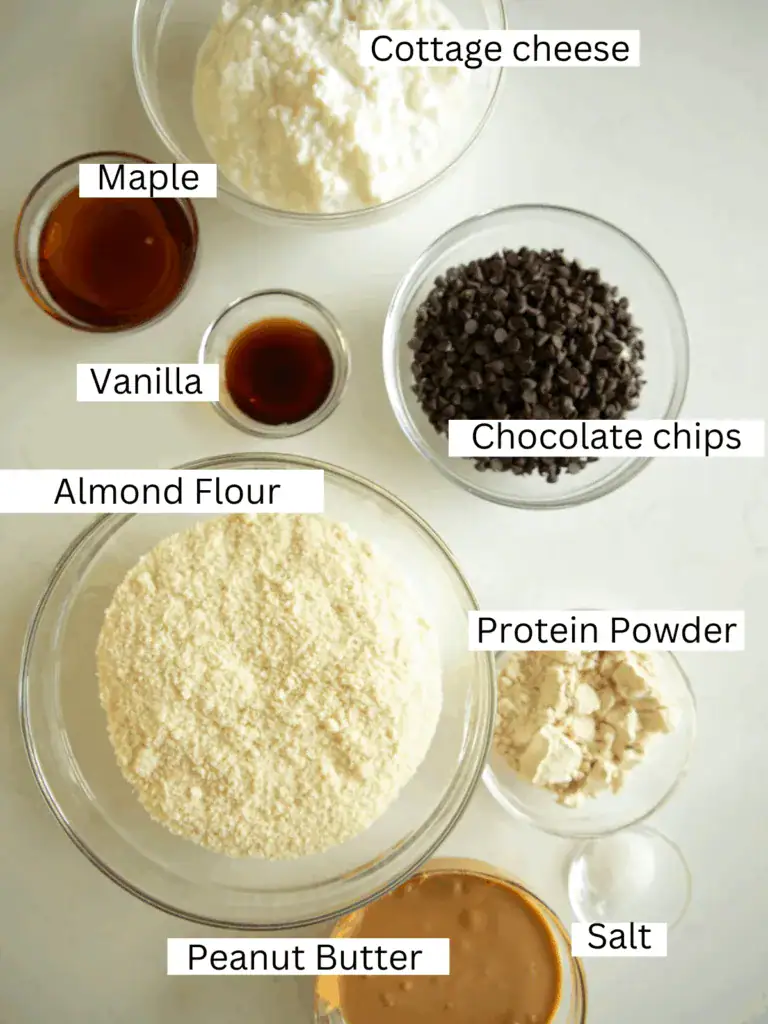 Overhead shot of labeled ingredients for healthy edible cookie dough: cottage cheese, peanut butter, almond flour, chocolate chips, protein powder, maple syrup, vanilla, and salt.