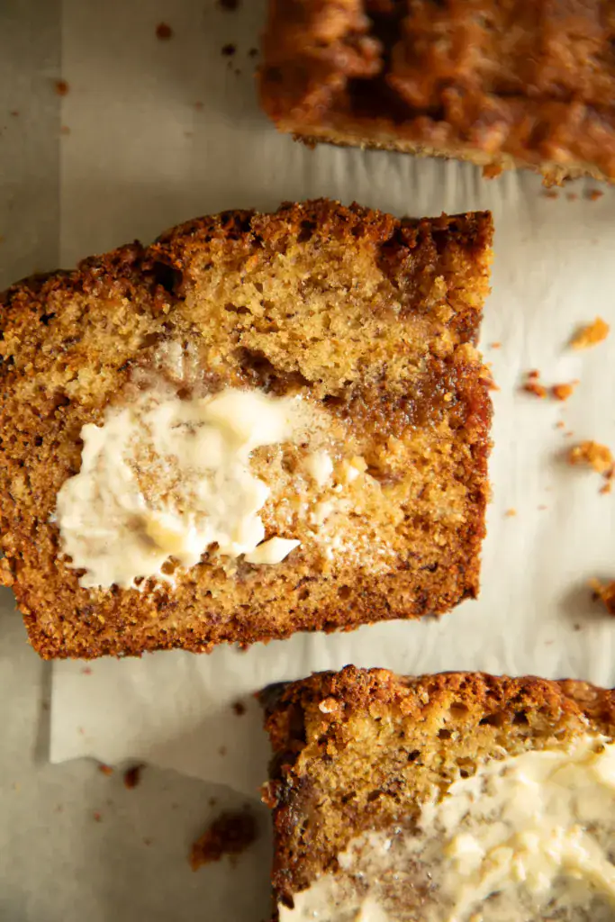 Sliced banana bread with a buttery top and full loaf in the background, both on parchment paper.