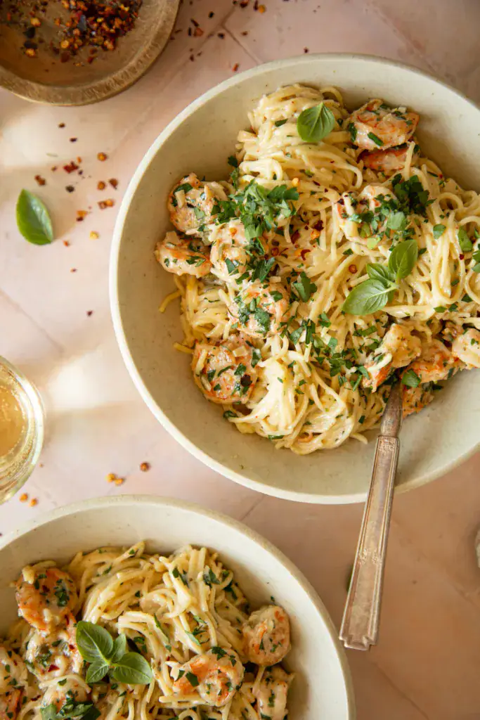 Multiple bowls of creamy shrimp scampi with parsley and basil on a pink tile surface.
