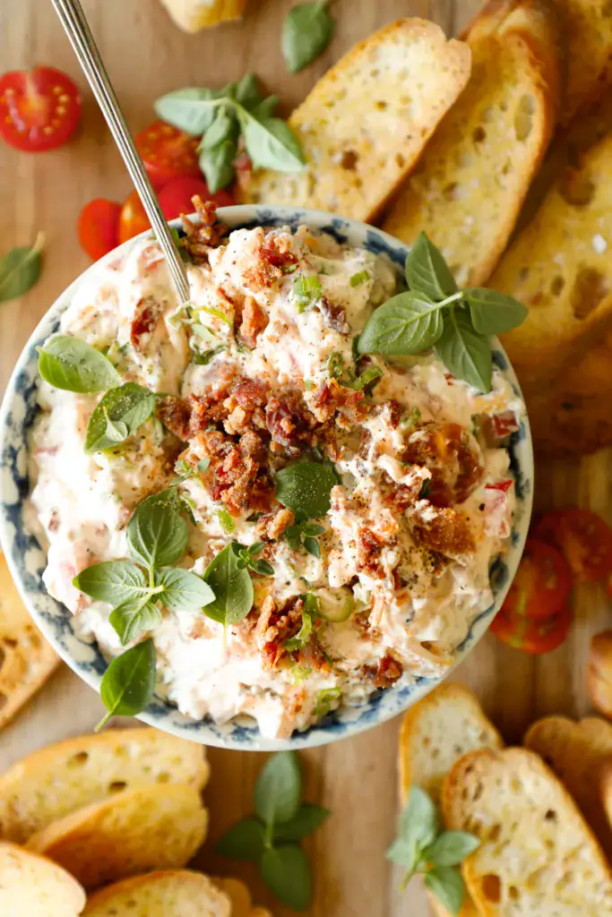 Overhead of BLT dip served with golden crostini, cherry tomatoes, and fresh basil on a wooden board