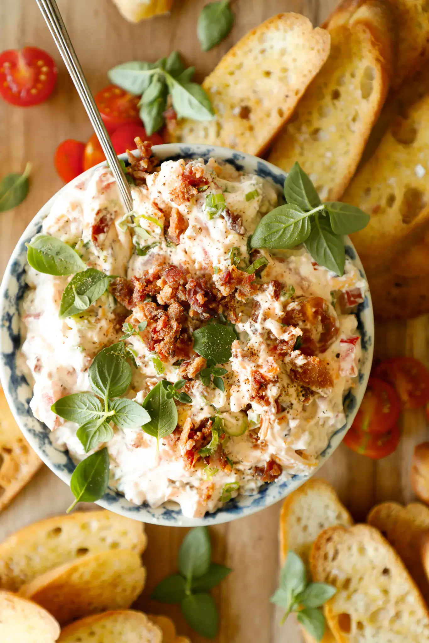 Overhead of BLT dip served with golden crostini, cherry tomatoes, and fresh basil on a wooden board