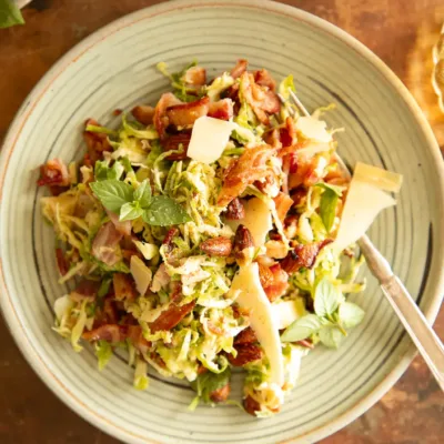 A serving of Brussels sprouts salad plated with bacon, shaved parmesan, almonds, and herbs on a green ceramic dish with a fork.