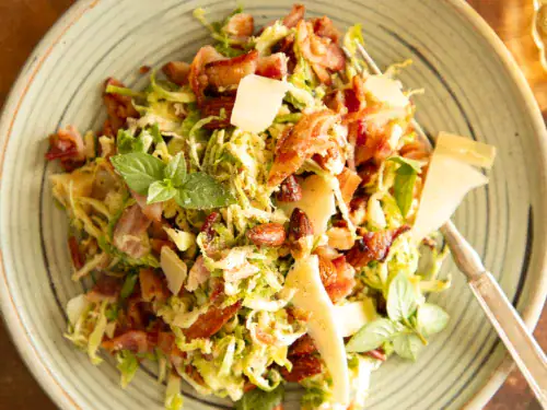 A serving of Brussels sprouts salad plated with bacon, shaved parmesan, almonds, and herbs on a green ceramic dish with a fork.