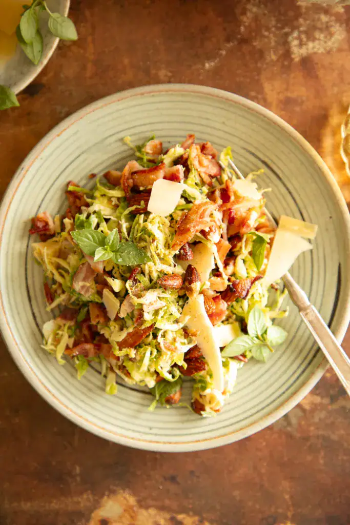 A serving of Brussels sprouts salad plated with bacon, shaved parmesan, almonds, and herbs on a green ceramic dish with a fork.