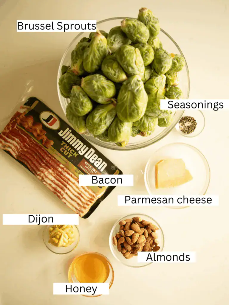 Overhead shot of raw Brussels sprouts, bacon, almonds, parmesan, honey, Dijon mustard, and seasonings arranged in bowls on a white surface.