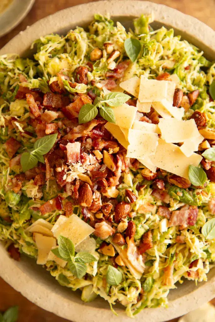 Close-up of shaved Brussels sprouts salad showing crispy bacon, golden almonds, parmesan shavings, and herbs.
