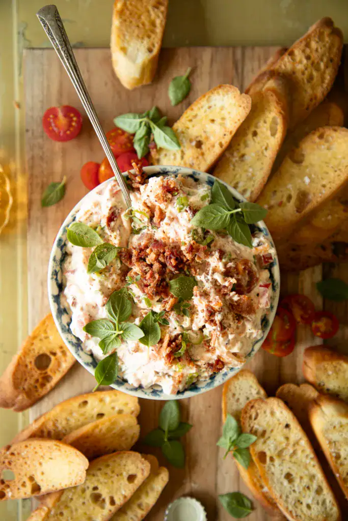 Bowl of creamy BLT dip topped with crispy bacon and fresh herbs, surrounded by toasted baguette slices