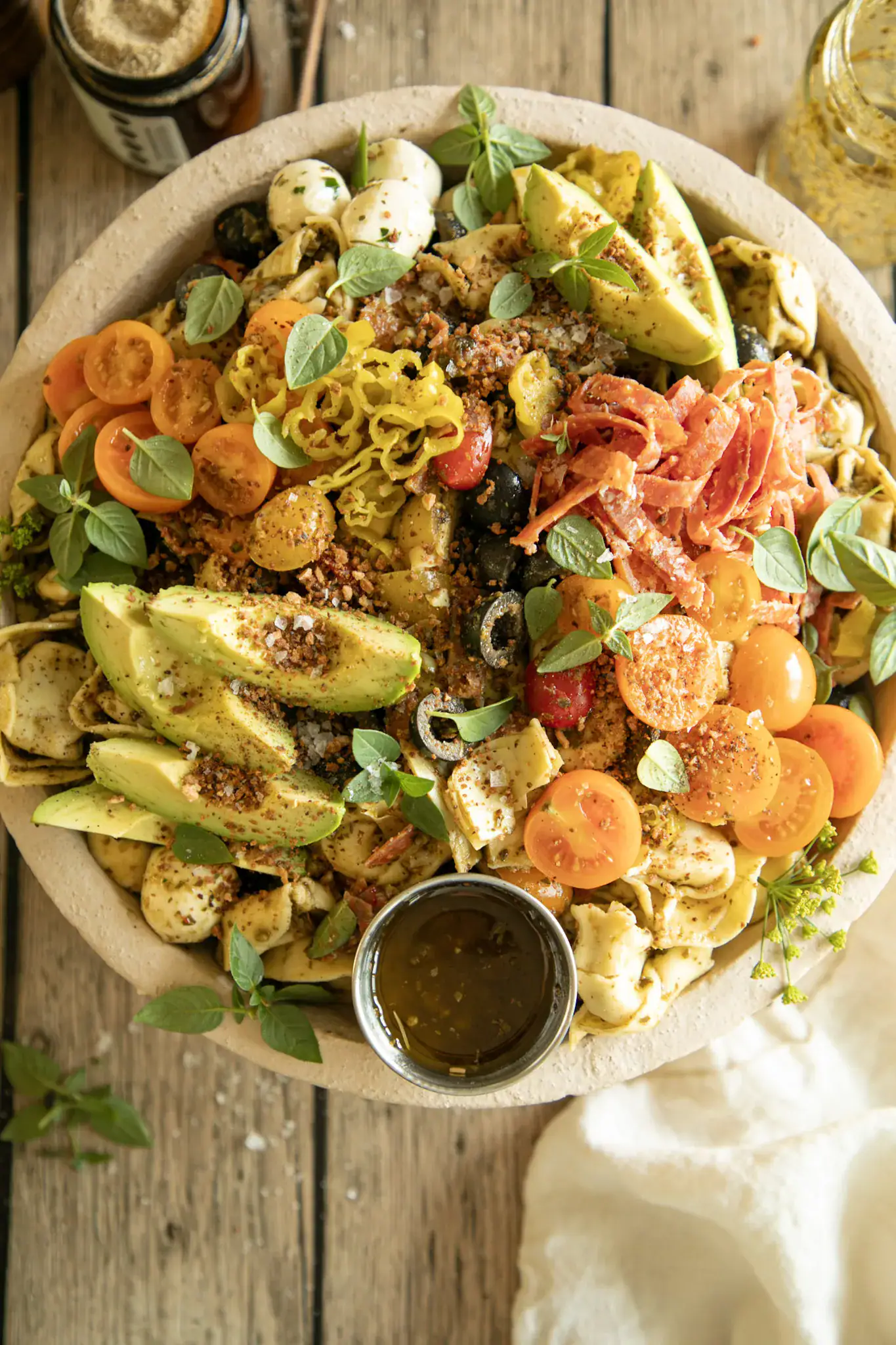 Overhead view of a rustic bowl filled with pesto tortellini salad and topped with tomatoes, salami, avocado, herbs, and a wooden spoon for serving.