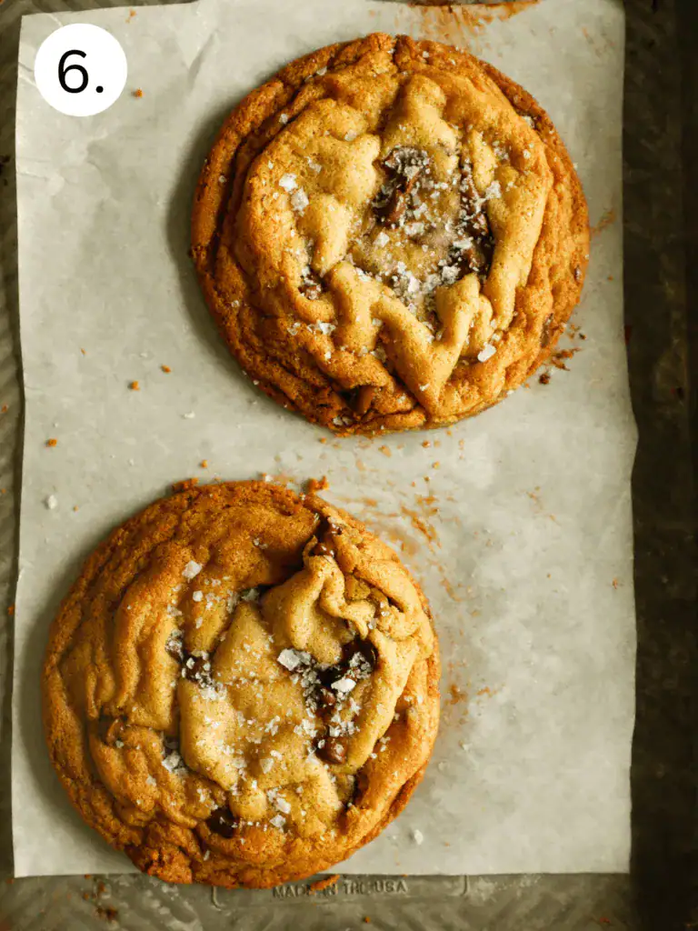 Two gooey chocolate chip cookies on a tray, surrounded by scattered chocolate chips and a small bowl of chips.