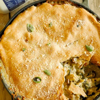 Overhead shot of whole baked chicken Alfredo pot pie in a cast iron skillet, garnished with Parmesan and herbs, next to AE Dairy milk.