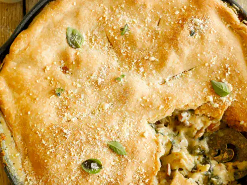 Overhead shot of whole baked chicken Alfredo pot pie in a cast iron skillet, garnished with Parmesan and herbs, next to AE Dairy milk.