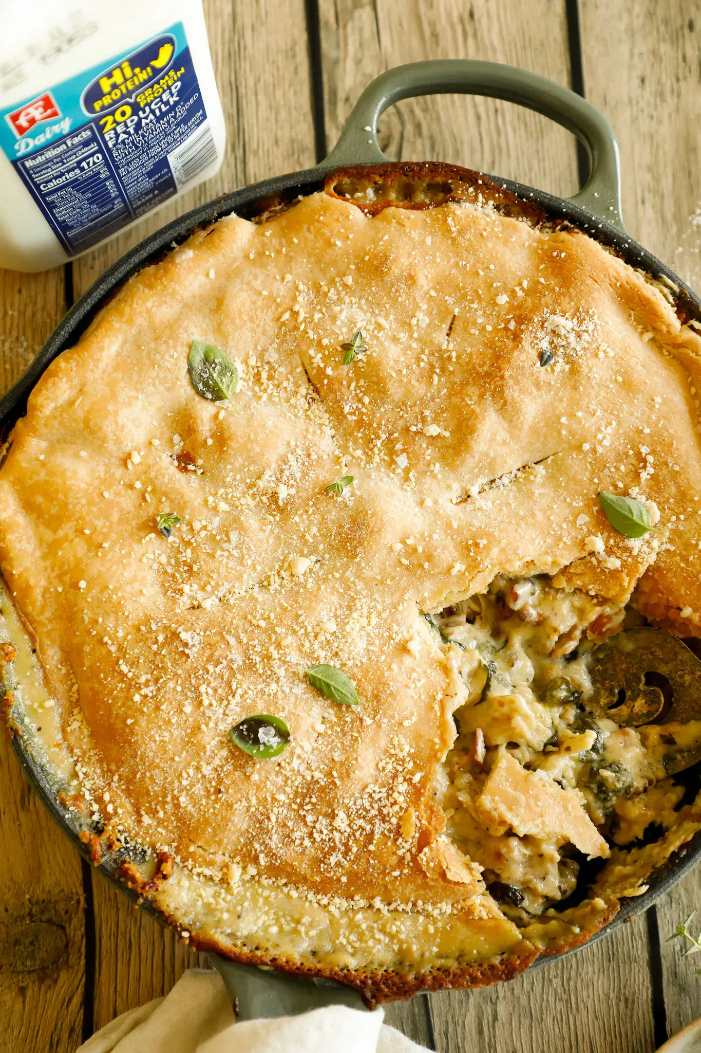 Overhead shot of whole baked chicken Alfredo pot pie in a cast iron skillet, garnished with Parmesan and herbs, next to AE Dairy milk.