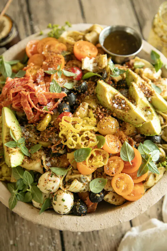 Full bowl of antipasto-style tortellini pasta salad served with a side of pesto dressing and fresh basil leaves.