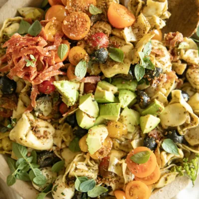 Bright, angled shot of tortellini pasta salad showing avocado slices, salami, tomatoes, pepperoncini, mozzarella balls, and olives, garnished with fresh herbs.