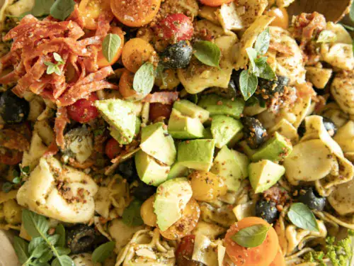 Bright, angled shot of tortellini pasta salad showing avocado slices, salami, tomatoes, pepperoncini, mozzarella balls, and olives, garnished with fresh herbs.