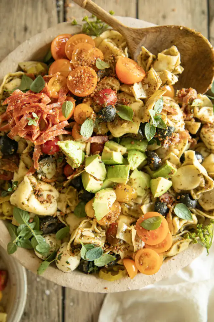 Bright, angled shot of tortellini pasta salad showing avocado slices, salami, tomatoes, pepperoncini, mozzarella balls, and olives, garnished with fresh herbs.
