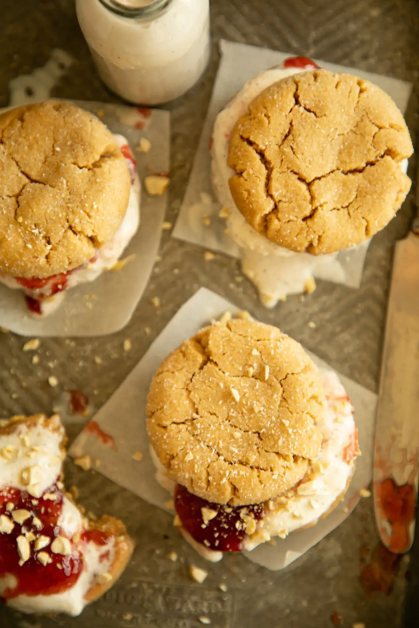 Flat lay of peanut butter cookies filled with strawberry jelly and vanilla ice cream, with a bottle of milk in the background