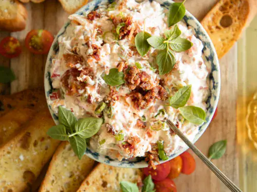 Bowl of creamy BLT dip topped with crispy bacon and fresh herbs, surrounded by toasted baguette slices