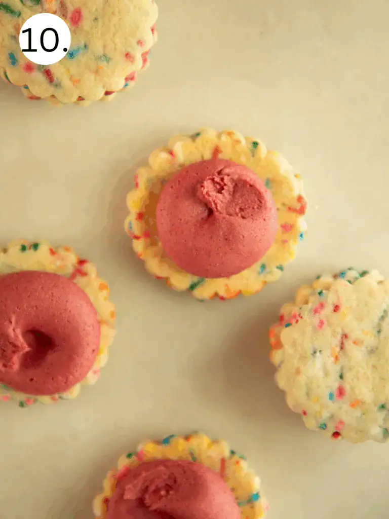 Partially assembled sandwich cookies with frosting piped on one side of the baked funfetti cookies.