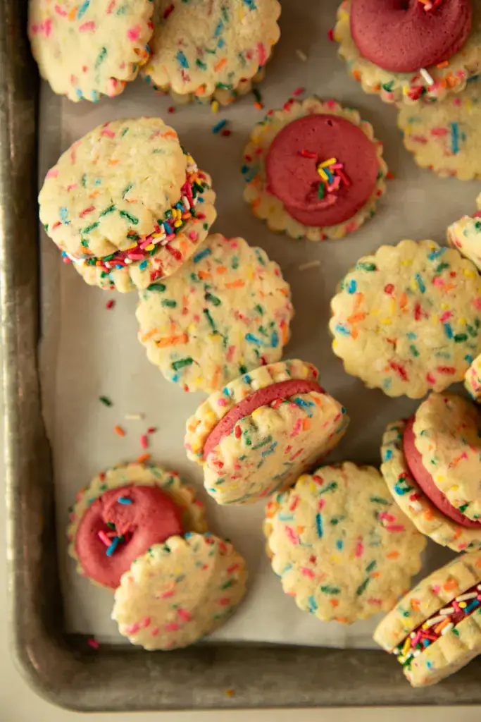 Overhead shot of funfetti shortbread cookie sandwiches filled with pink frosting and rolled in sprinkles.