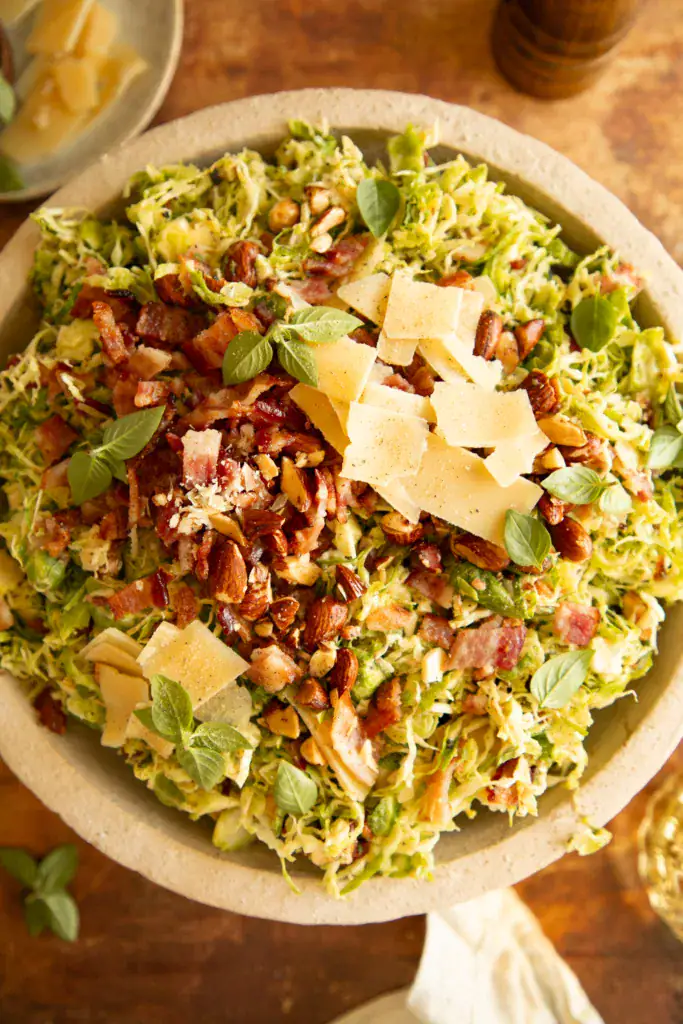 Overhead shot of Brussels sprout salad in a stone bowl, topped with chopped bacon, shaved parmesan, toasted almonds, and fresh herbs.